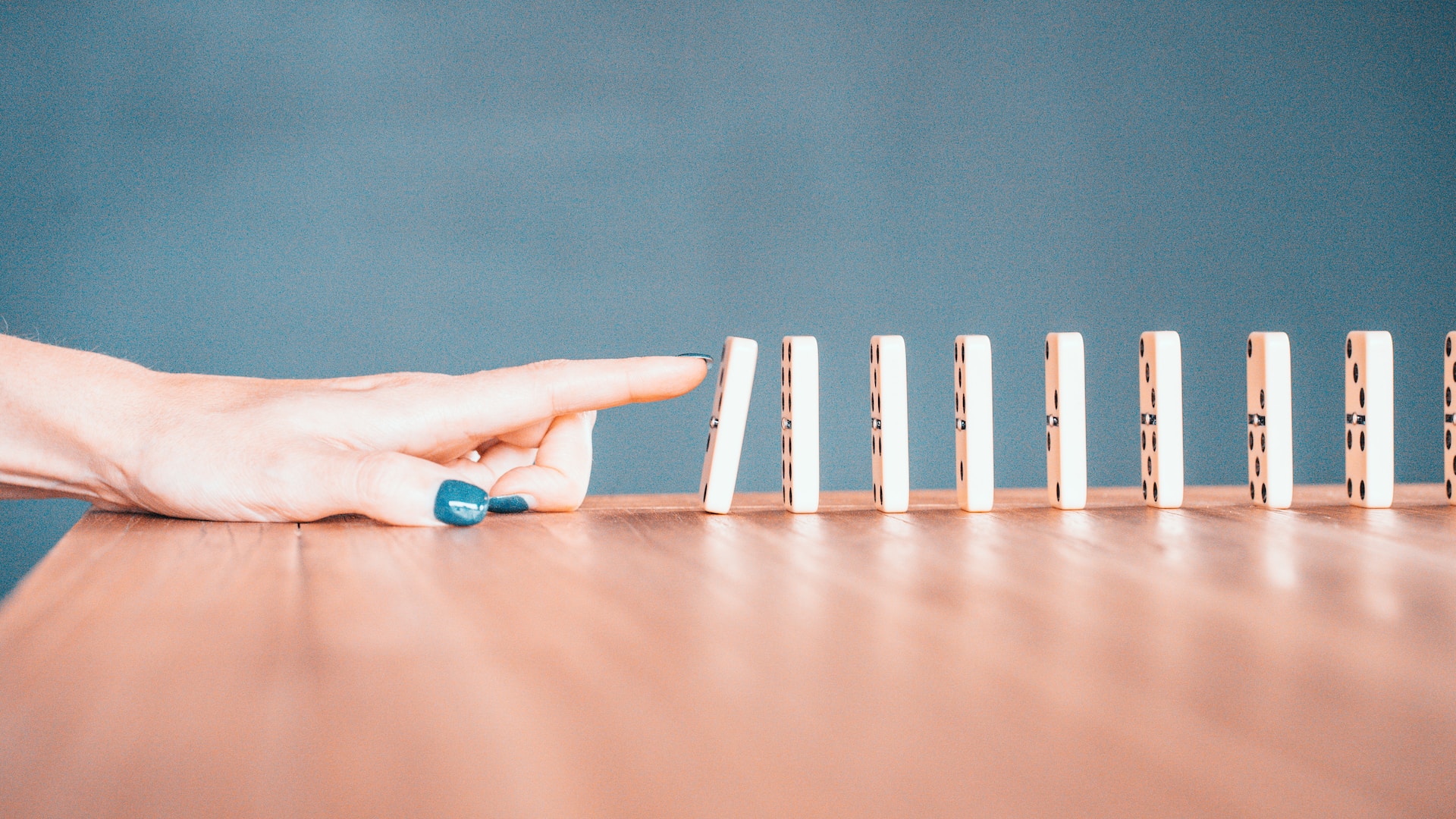 description: Hand, with finger extended, tipping over the first in a line of dominoes. Photo by Bradyn Trollip on Unsplash