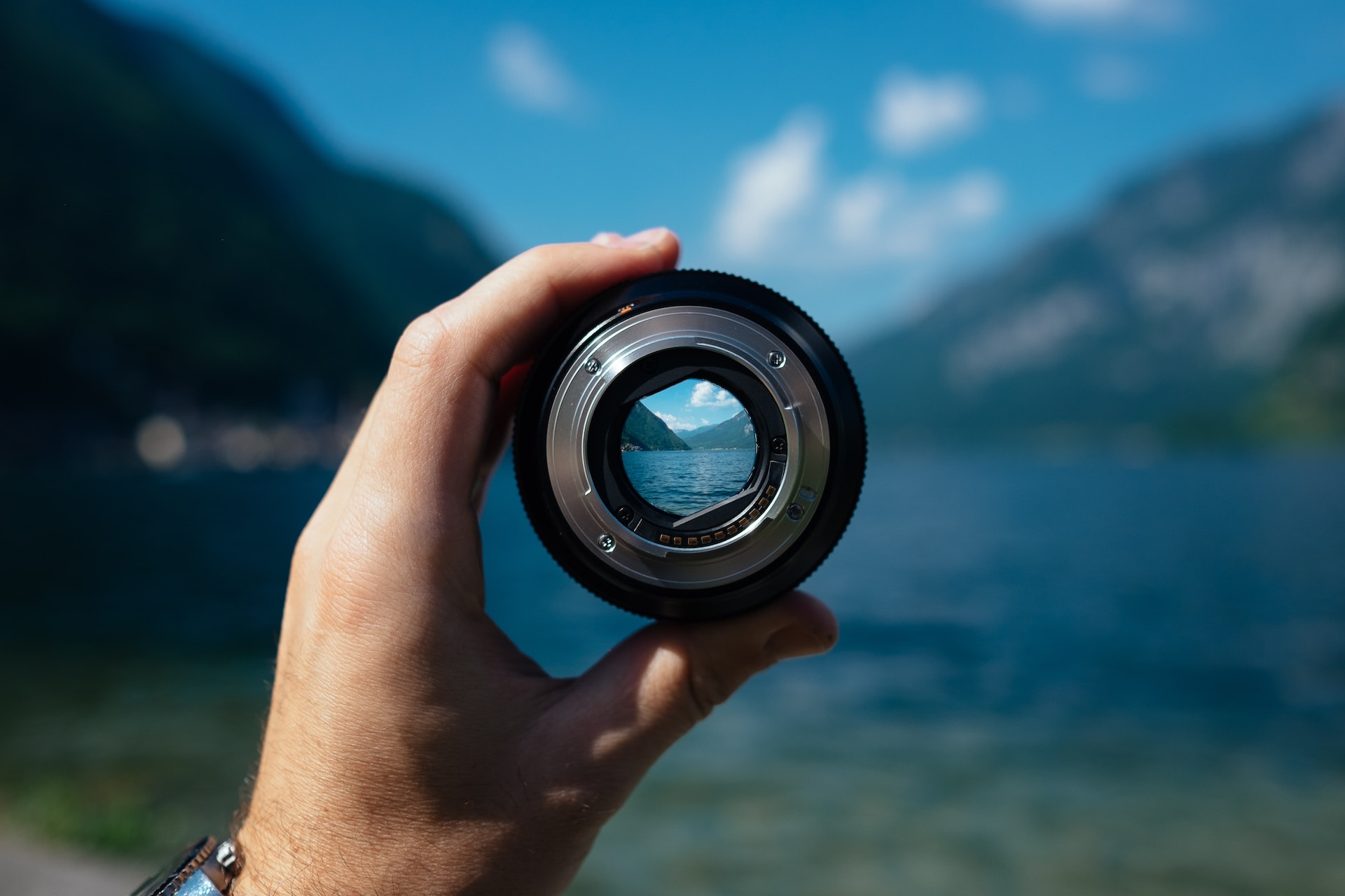 A hand holding a camera lens in front of a mountain lake scene.  The image outside of the lens is blurry; inside the lens, it is clear and focused.