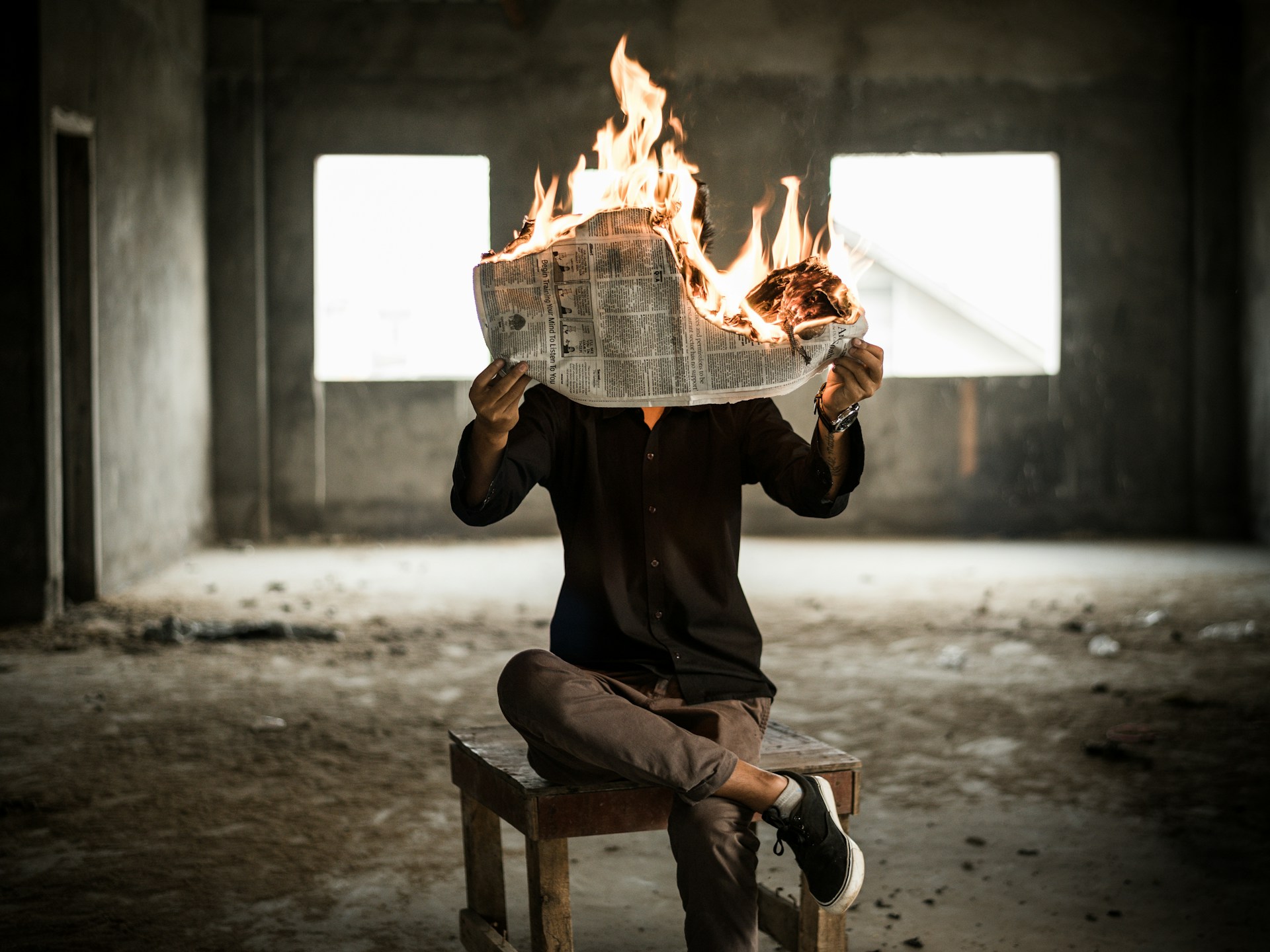 An image of a person, appearing to read a newspaper that is on fire.  They are sitting in an empty room.Photo by Nijwam Swargiary on Unsplash