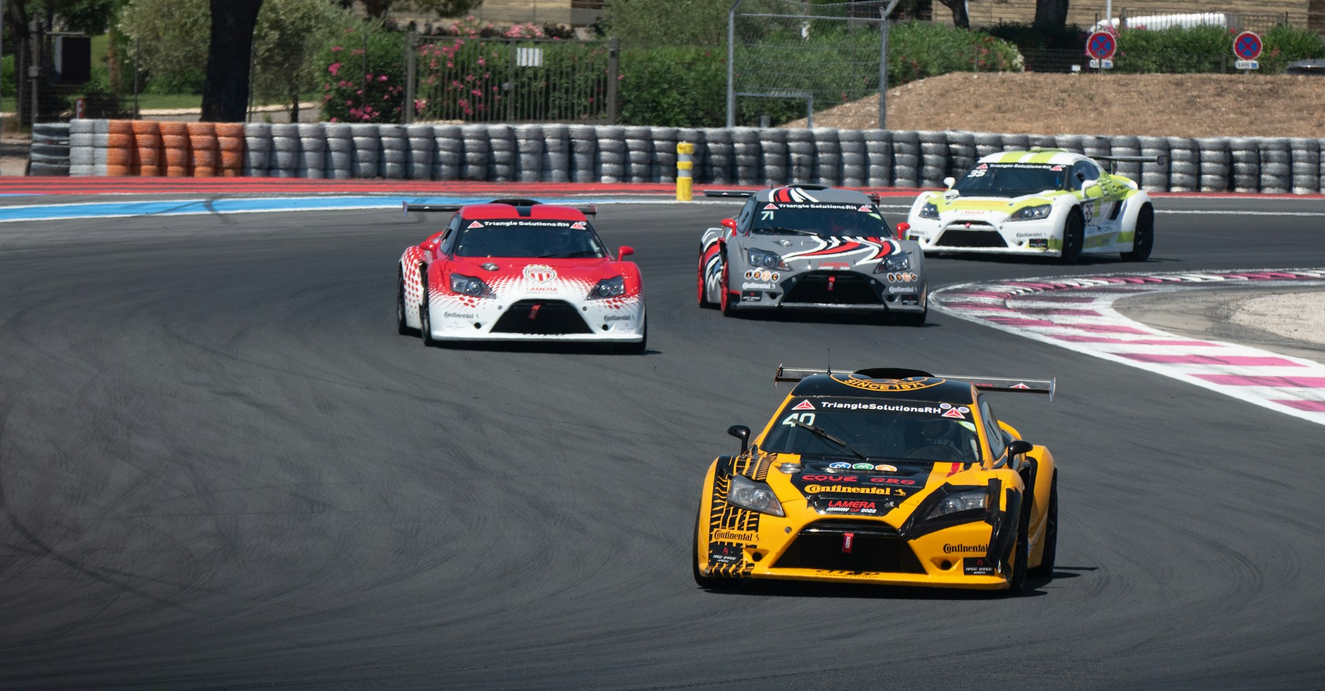 Race cars on a track.  A yellow car leads, with three other cars trailing behind it.  Photo by Christopher John on Unsplash.