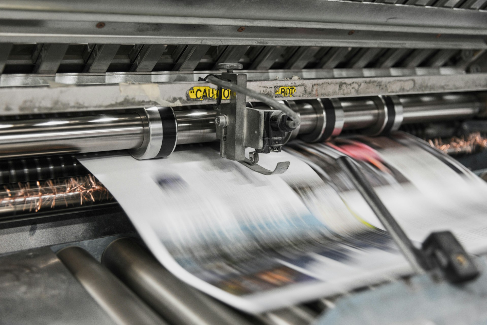 A newspaper printing machine in action. The paper, which is in motion, is a blur.  Photo by Bank Phrom on Unsplash.