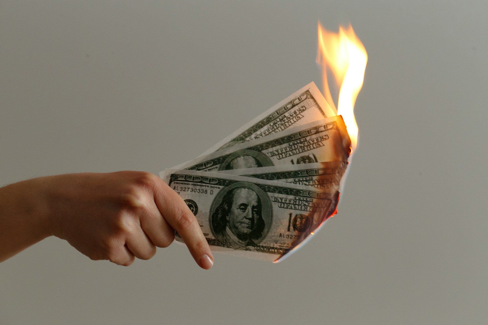 a hand holds up three US $100 bills that are on fire. Photo by Jp Valery on Unsplash.